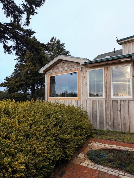 Wooden building with windows, a brick path, and a large green bush under an overcast sky.