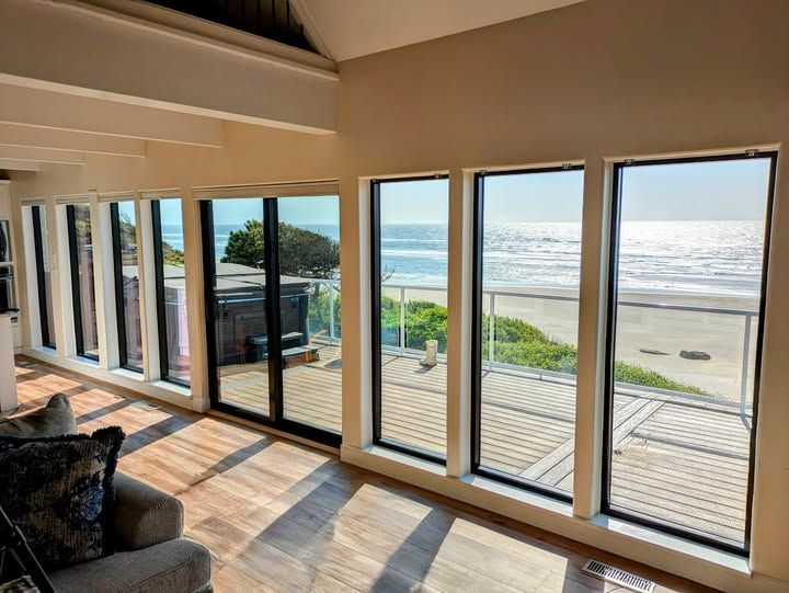Interior view with windows facing the ocean. Beach and sunlight visible, wood floors, neutral walls, and seating.