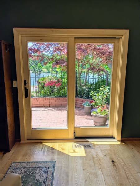 Sliding glass doors open onto a patio with red brick, flowers, and greenery. Sunlight streams into the room.