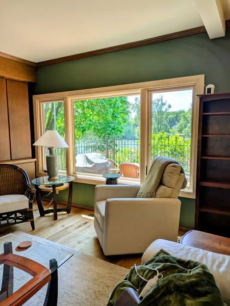 Cozy living room with large windows overlooking trees. White armchair, green wall, wooden floors.