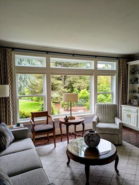 Living room with large windows overlooking a green yard; furnished with a sofa, chairs, and a round coffee table.
