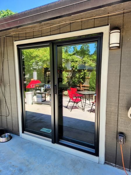 Sliding glass doors with black frames reflect a yard with a red chair and trees, set in a light-colored frame.