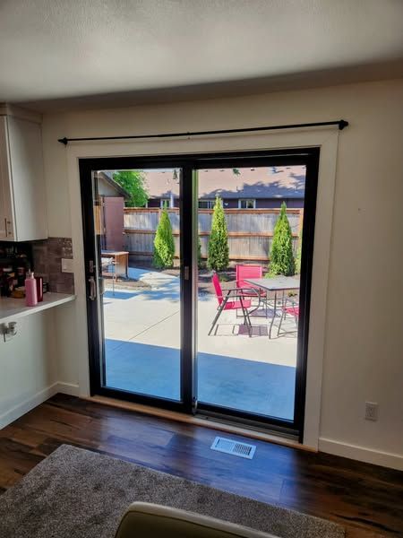 Sliding glass doors with black frames open to a patio with red chairs and green trees; inside, wood floors and white walls.