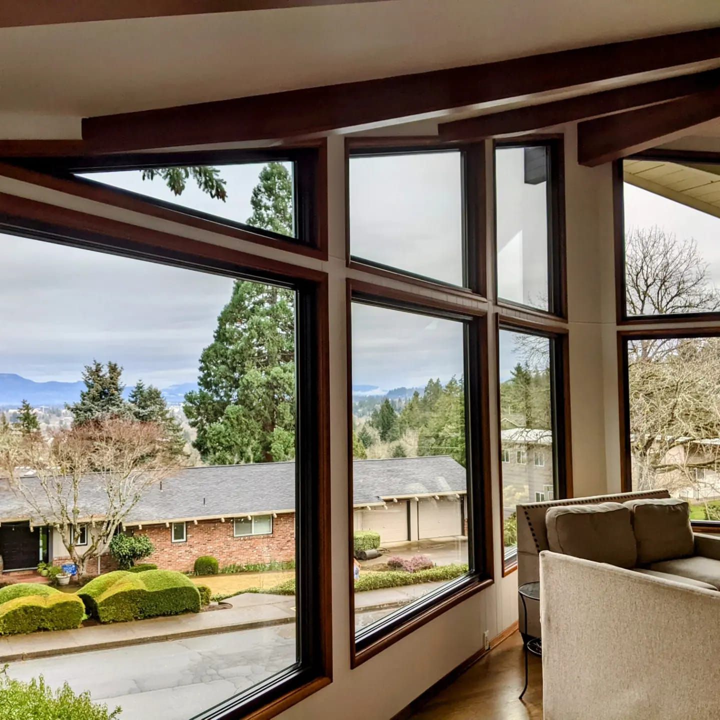 Large windows with a view of houses, trees, and hills. Brown window frames, cream ceiling, and a beige sofa.