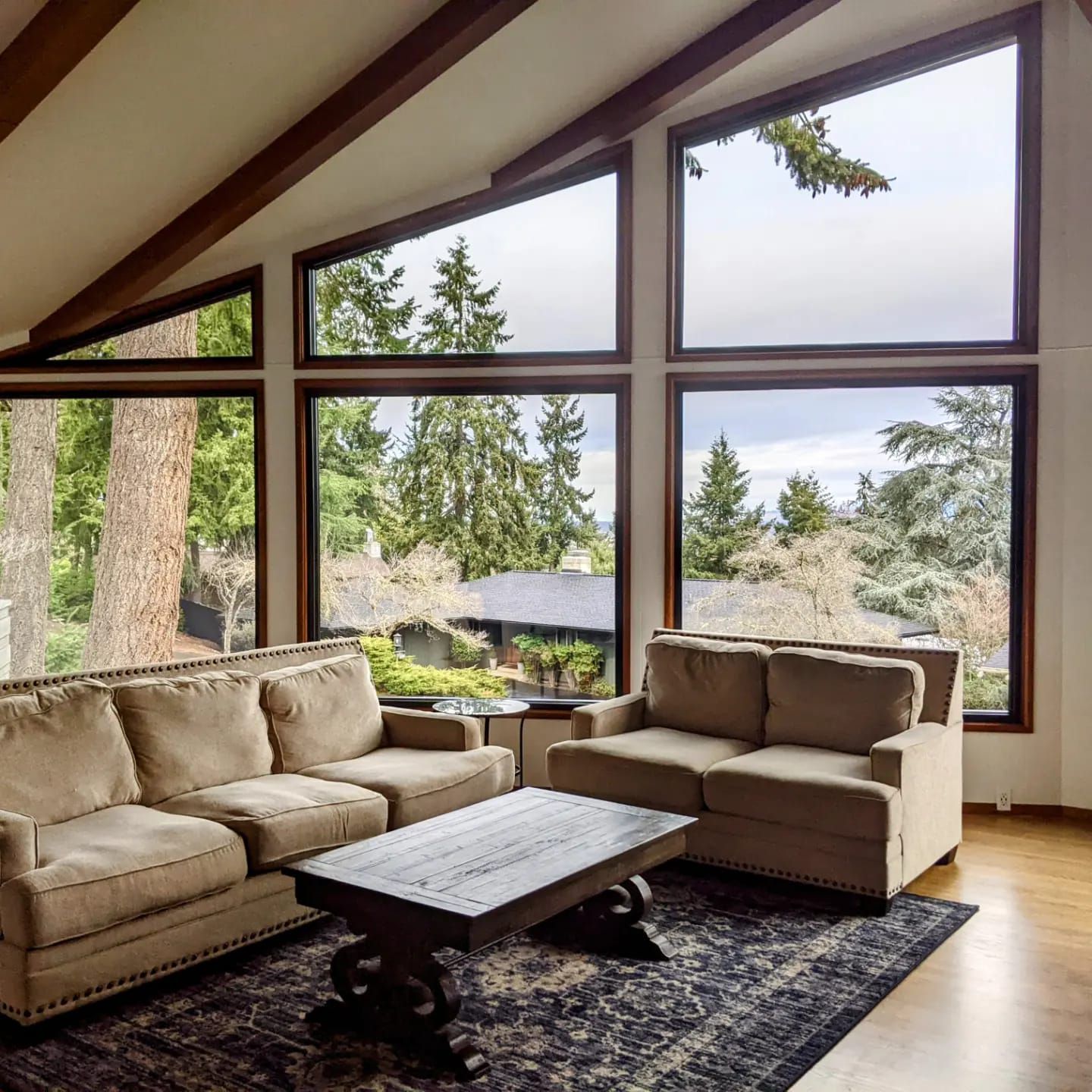 Living room with large windows, light brown couches, dark rug, and wood coffee table.