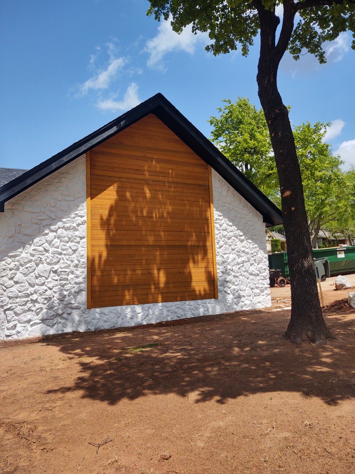 White building with a wooden gable panel and dark roof under a blue sky, beside a tree on red dirt.