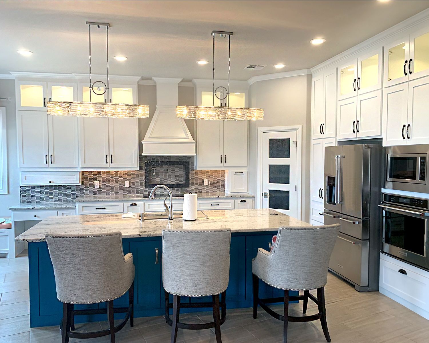 Modern white kitchen with marble island, three chairs, pendant lights, stainless appliances, and built-in oven