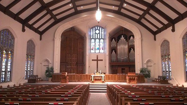 Interior of a church with wooden pews, altar, and organ. Large stained-glass window at the back.