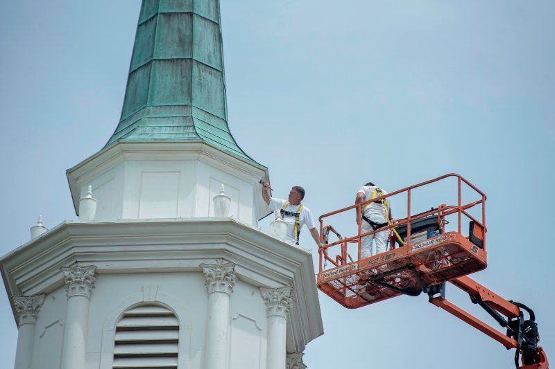 Two workers on a lift painting the white spire of a building with a copper-colored top against a blue sky.