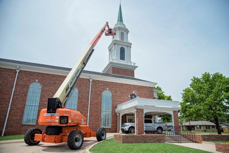 An orange boom lift extending to the steeple of a brick church; a white truck is parked under the covered entrance.