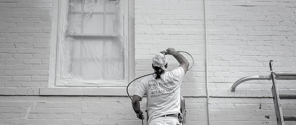 Person painting a brick building with a paint sprayer, window taped off, monochrome.