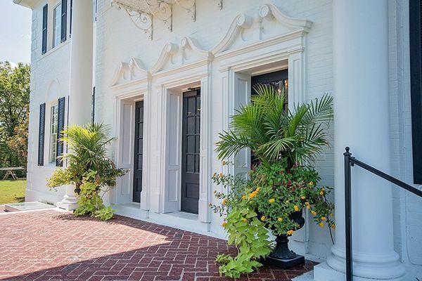 White building facade with black doors, decorative trim, potted plants, and red brick pathway.