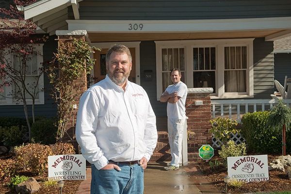 Two men in front of a house; one in a white shirt and jeans, the other behind him. Banners read 