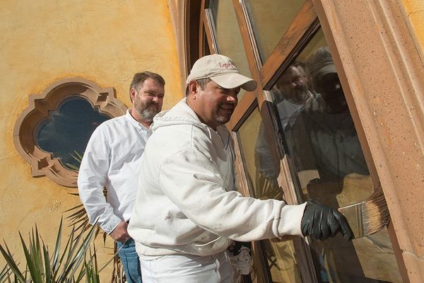 Two men painting window trim of a building with ornate facade; one paints, the other watches.