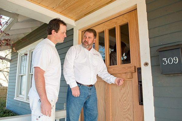 Two men at front door; one points, other listens. Door is wood; house is blue.