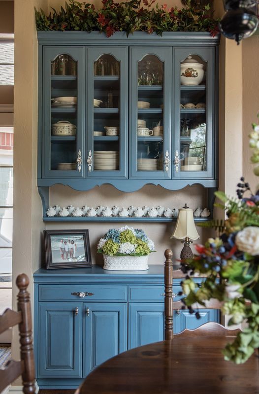 Blue hutch with glass-fronted cabinets displaying dishware; topped with greenery, next to a table setting.