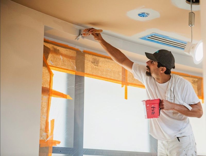 A person paints a ceiling with a brush, holding a paint can. Window masked with orange.