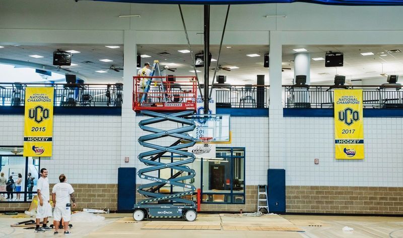 People working on a scissor lift in a gymnasium, banners on walls, equipment on the floor.