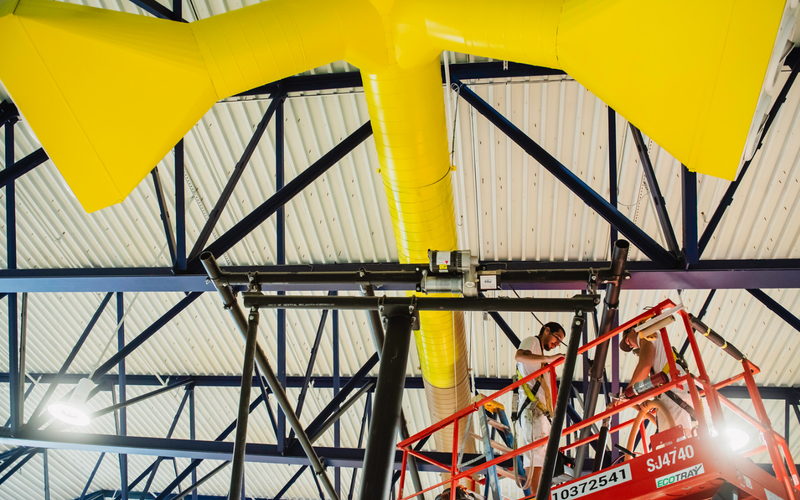 Two workers on a lift installing a large yellow air duct within a metal-framed structure.