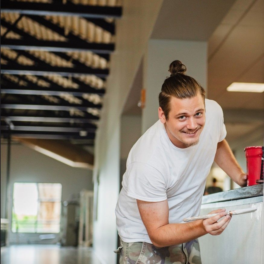 Man with topknot smiles, holding a paintbrush and red cup, paints in a hallway.