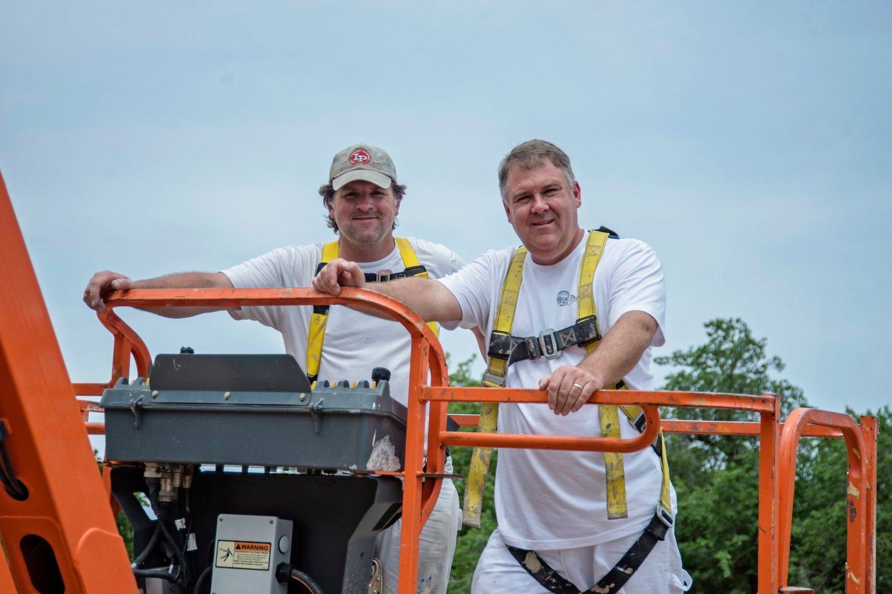 Two men on an orange lift wearing harnesses, smiling at the camera, against a cloudy sky.