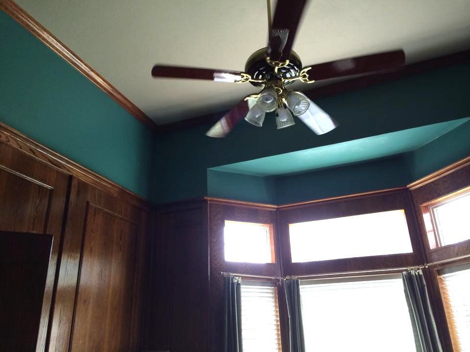 Ceiling fan in a room with dark teal walls, wood trim, and a bay window.