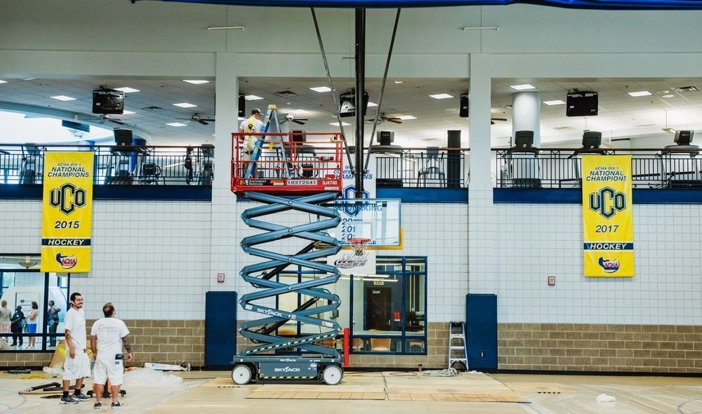 Workers in a scissor lift working on a basketball hoop inside a gymnasium. Banners hang from the ceiling.