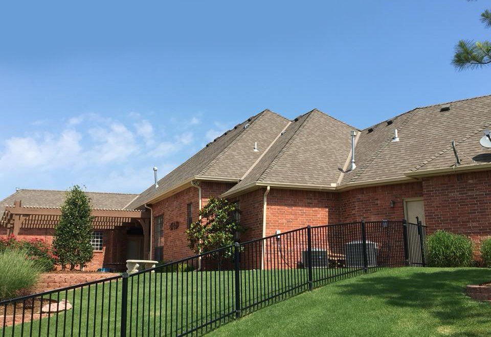 Red brick house with a brown shingled roof, black fence, and green lawn against a blue sky.