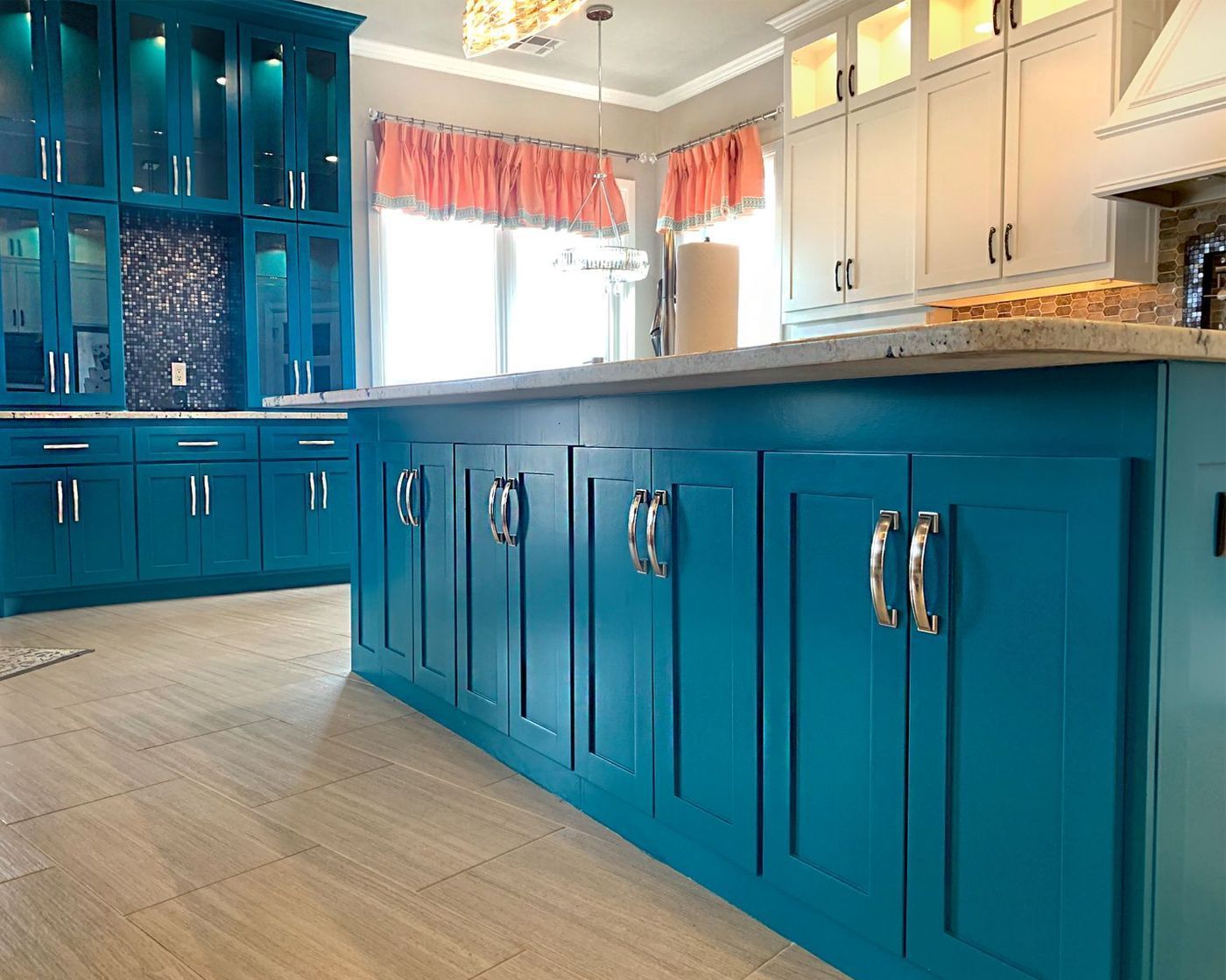 Turquoise kitchen island and cabinets, light wood floor, white and cream upper cabinets, window with pink valance.