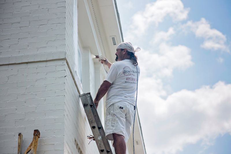 Man on ladder painting white trim of a white building, against a partly cloudy sky.