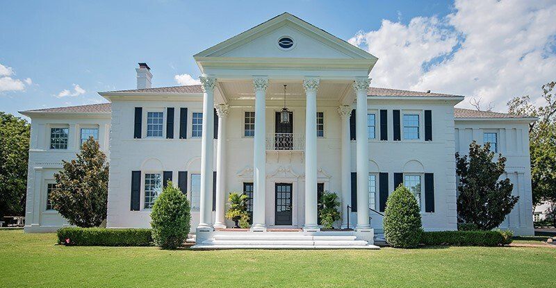 White mansion with Greek columns, black shutters, and a green lawn under a blue sky.