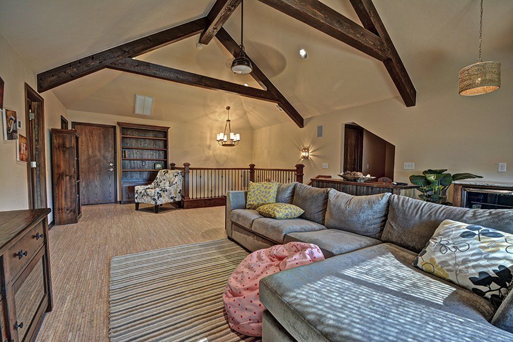 Cozy living room with gray sectional sofa, exposed beams, and a rug. Bookshelves and a doorway in the background.