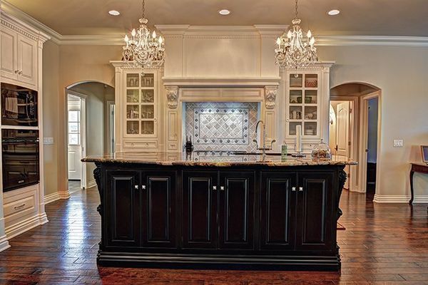 Elegant kitchen with dark island and light cabinets; two crystal chandeliers hang above.