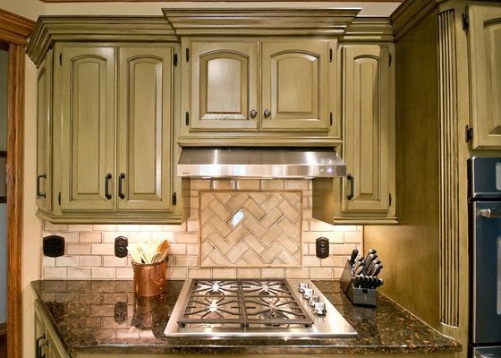 Kitchen cabinets in shades of green, above a stainless steel stovetop and tan backsplash.