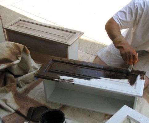 Person staining a white cabinet door brown with a brush; other cabinet pieces are nearby.