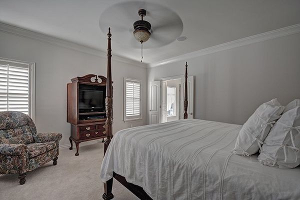 Bedroom with four-poster bed, armchair, TV cabinet, shuttered windows, and a ceiling fan.