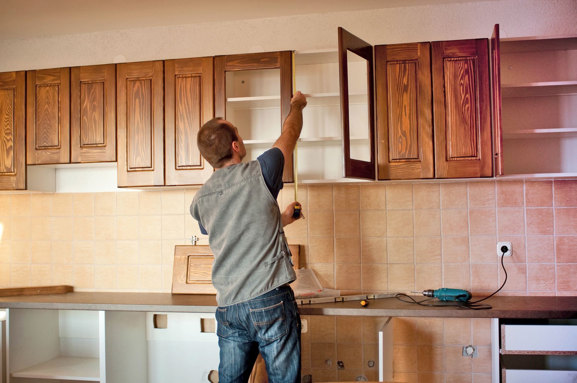 Person measuring cabinet in a kitchen during renovation, wooden cabinets, tools on counter.