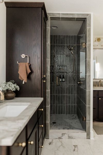 Bathroom with dark cabinets, marble countertop, and a walk-in shower with vertical tile.