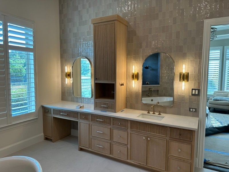Bathroom with light wood cabinets, two arched mirrors, gold sconces, and a window with shutters.