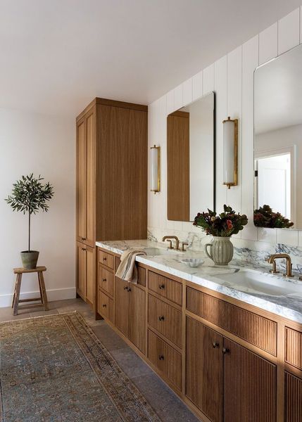 Bathroom with wood cabinetry, marble countertops, and a patterned rug. A small tree sits on a stool.