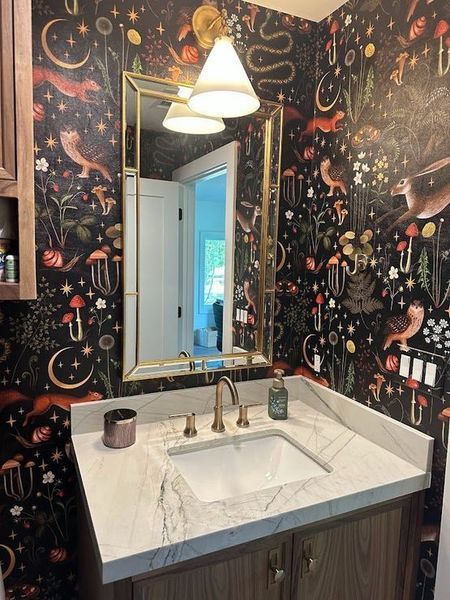 Bathroom with whimsical wallpaper, gold-framed mirror, white sink, and light fixtures.