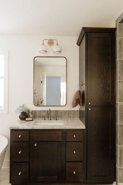 Bathroom with dark wood vanity, tall cabinet, gold-framed mirror, and two-light fixture.