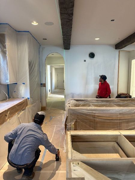 Two workers paint a room under construction, one kneeling, one standing, with partially finished cabinets and exposed beams.