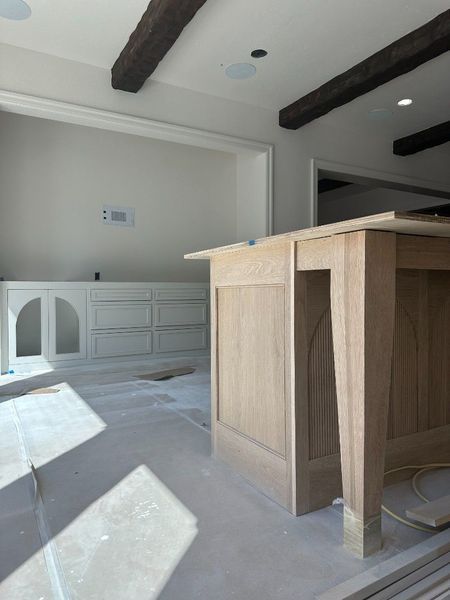Kitchen under construction with unfinished light-wood island, cabinets, dark beams, and bright natural light.