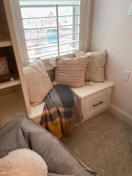 Cozy window seat with pillows and a blanket. Built-in storage, light-colored walls and carpet.