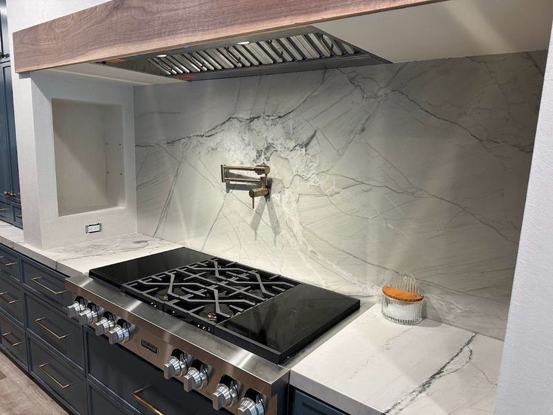 Kitchen with a marble backsplash, stainless steel range, and dark blue cabinets. A wooden range hood is present.