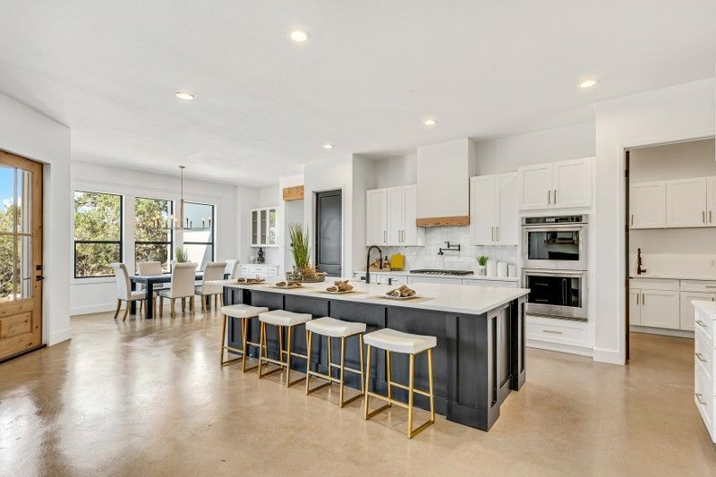 Open-concept kitchen with large island, seating, and white cabinets. Dining table, windows, and light wood door.