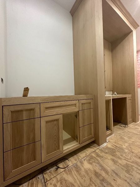Unfinished wooden bathroom cabinets and built-in desk under construction against a white wall and unfinished wood floor.