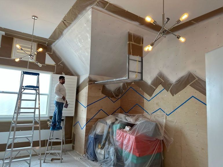 Man on ladder painting wall during interior renovation; blue tape detail, brown paper protective covering, overhead light fixtures.