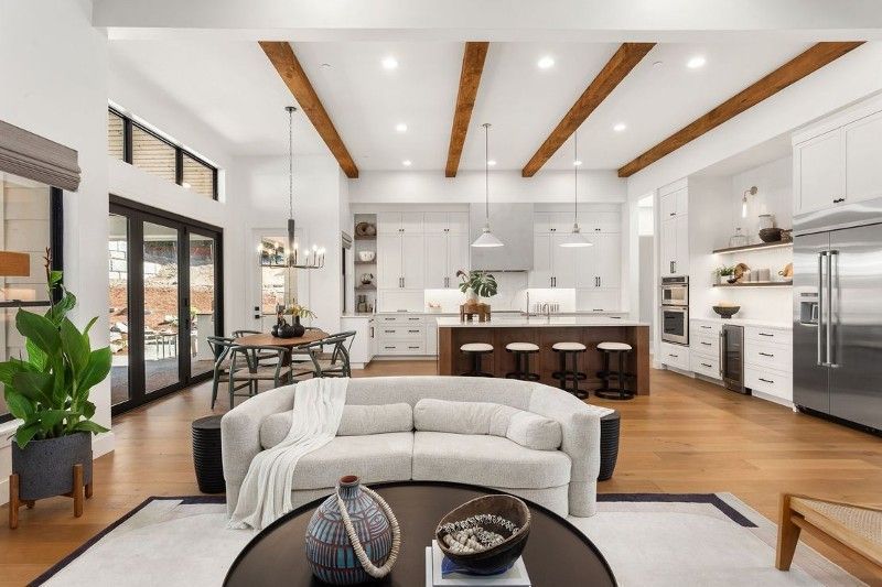 Open-plan living space with kitchen, dining, and living areas. White and wood tones, large windows, and modern furniture.